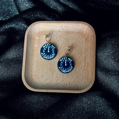 Round black earrings with midnight clock detailing and "Meet Me At Midnight" text, resting in a wooden tray on sparkling black fabric. 