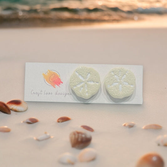 Close-up of handmade polymer clay sand dollar earrings on a beach-themed background with seashells, perfect for ocean lovers. 