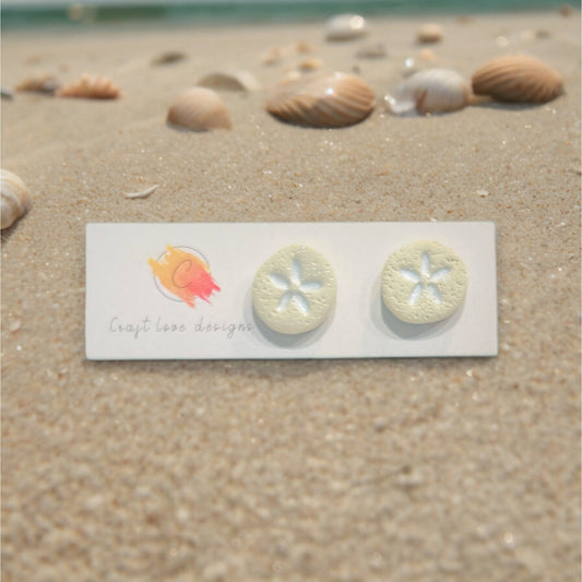 Polymer clay sand dollar earrings displayed on a product card, set against a coastal backdrop with shells scattered on warm sand. 