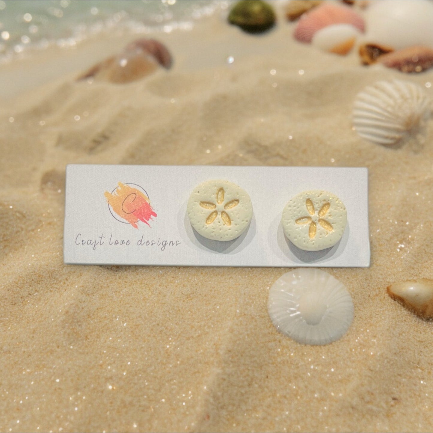 Handmade polymer clay sand dollar stud earrings displayed on a beach backdrop with shells and sand for a coastal vibe.