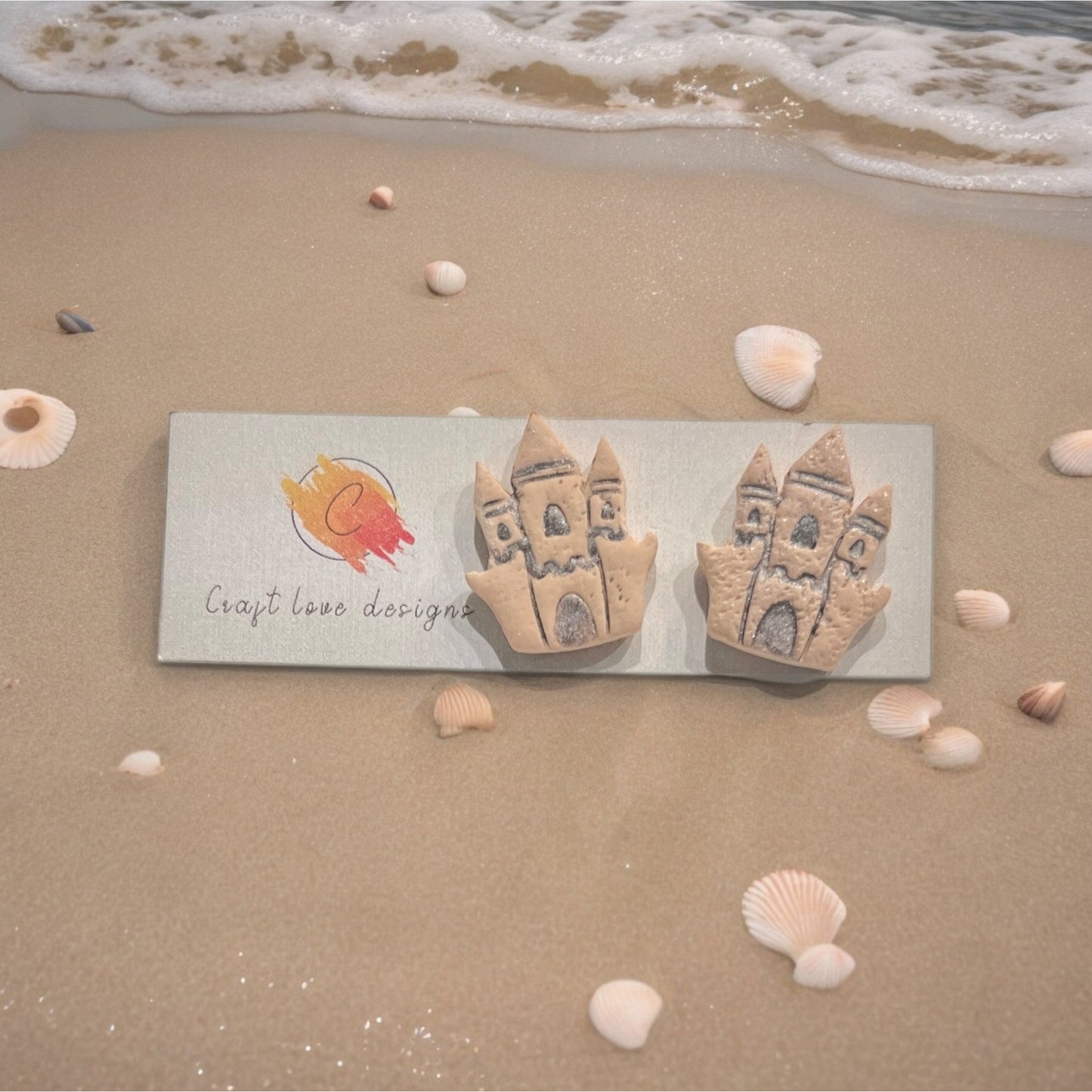 Handmade sandcastle-themed earrings placed on a Craft Love Designs logo card, surrounded by seashells and gentle ocean waves.