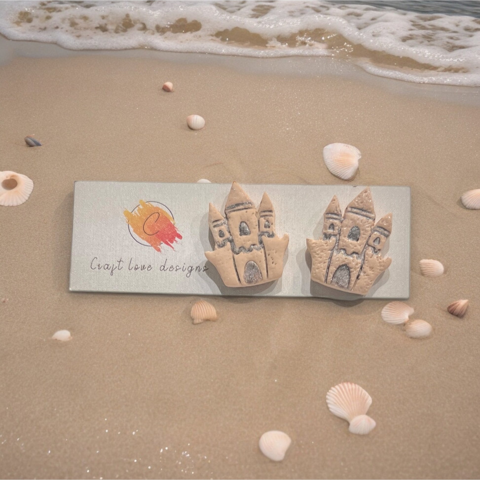 Handmade sandcastle-themed earrings placed on a Craft Love Designs logo card, surrounded by seashells and gentle ocean waves.