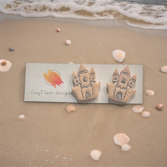 Handmade sandcastle-themed earrings placed on a Craft Love Designs logo card, surrounded by seashells and gentle ocean waves.