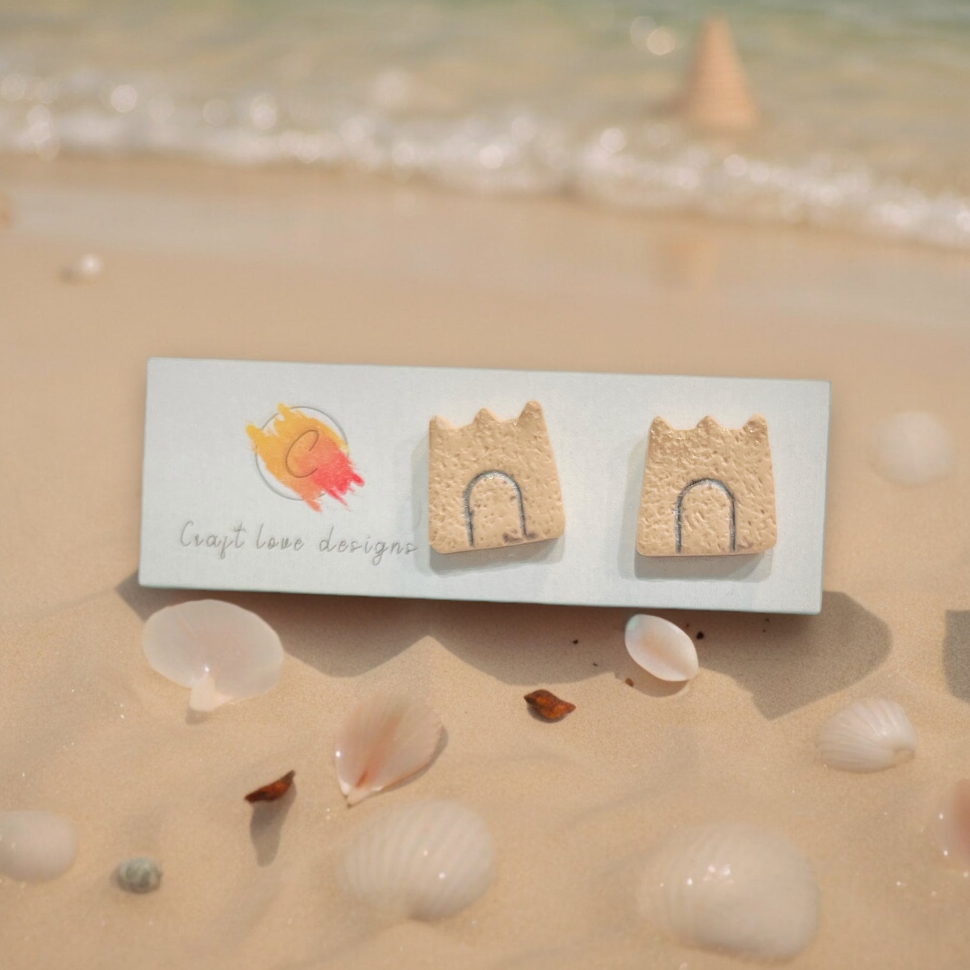 Sandcastle stud earrings displayed on a beach-themed card with seashells scattered on the sand.