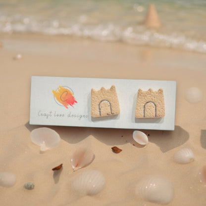 Sandcastle stud earrings displayed on a beach-themed card with seashells scattered on the sand.