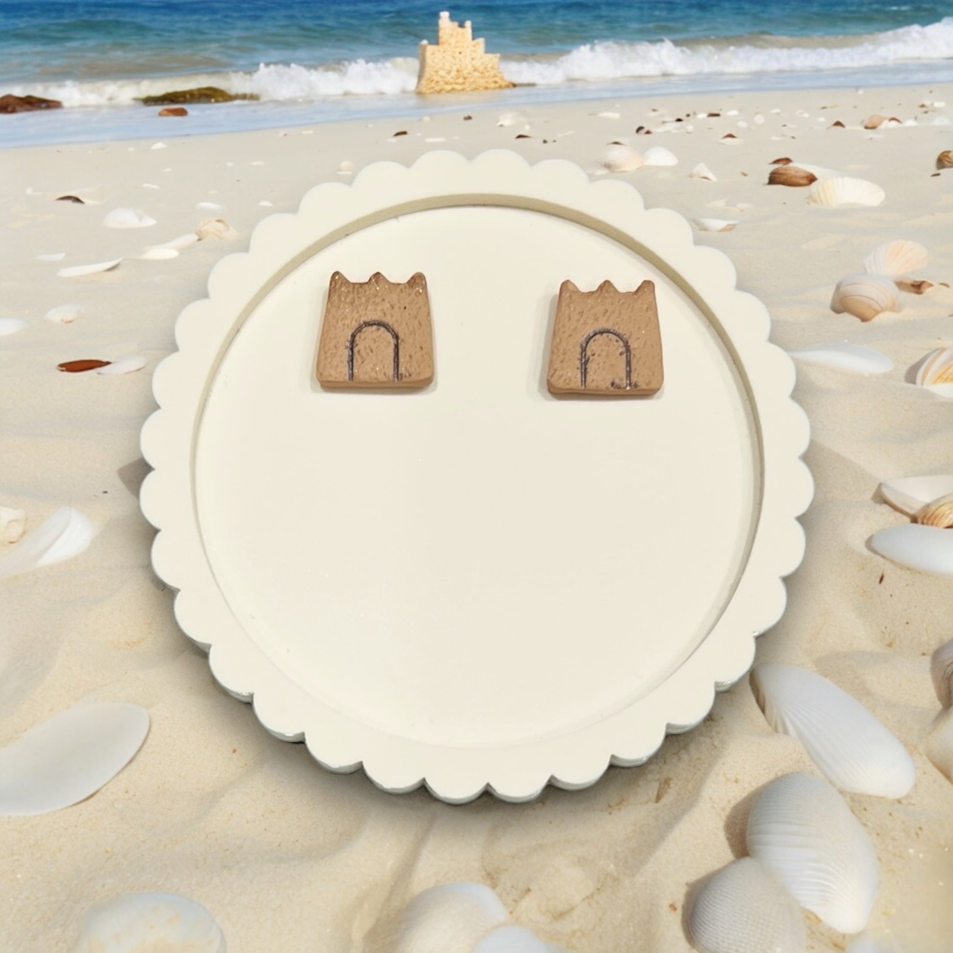 Sandcastle earrings resting on a decorative round white tray, surrounded by seashells and sand.