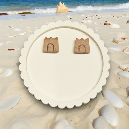 Sandcastle earrings resting on a decorative round white tray, surrounded by seashells and sand.