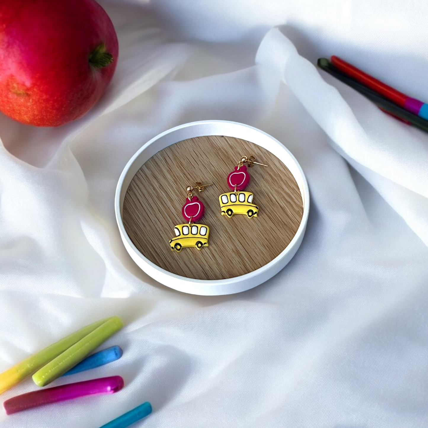 Handmade school bus and apple earrings placed in a wooden dish, surrounded by colorful crayons and red apples over soft white fabric.