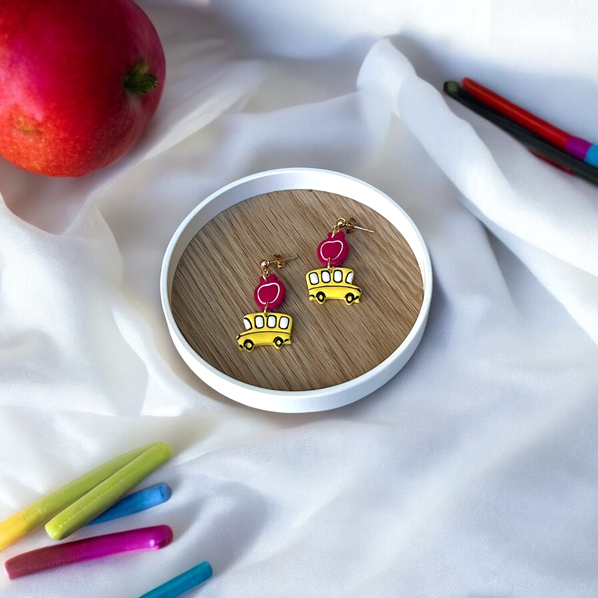 Handmade school bus and apple earrings placed in a wooden dish, surrounded by colorful crayons and red apples over soft white fabric.