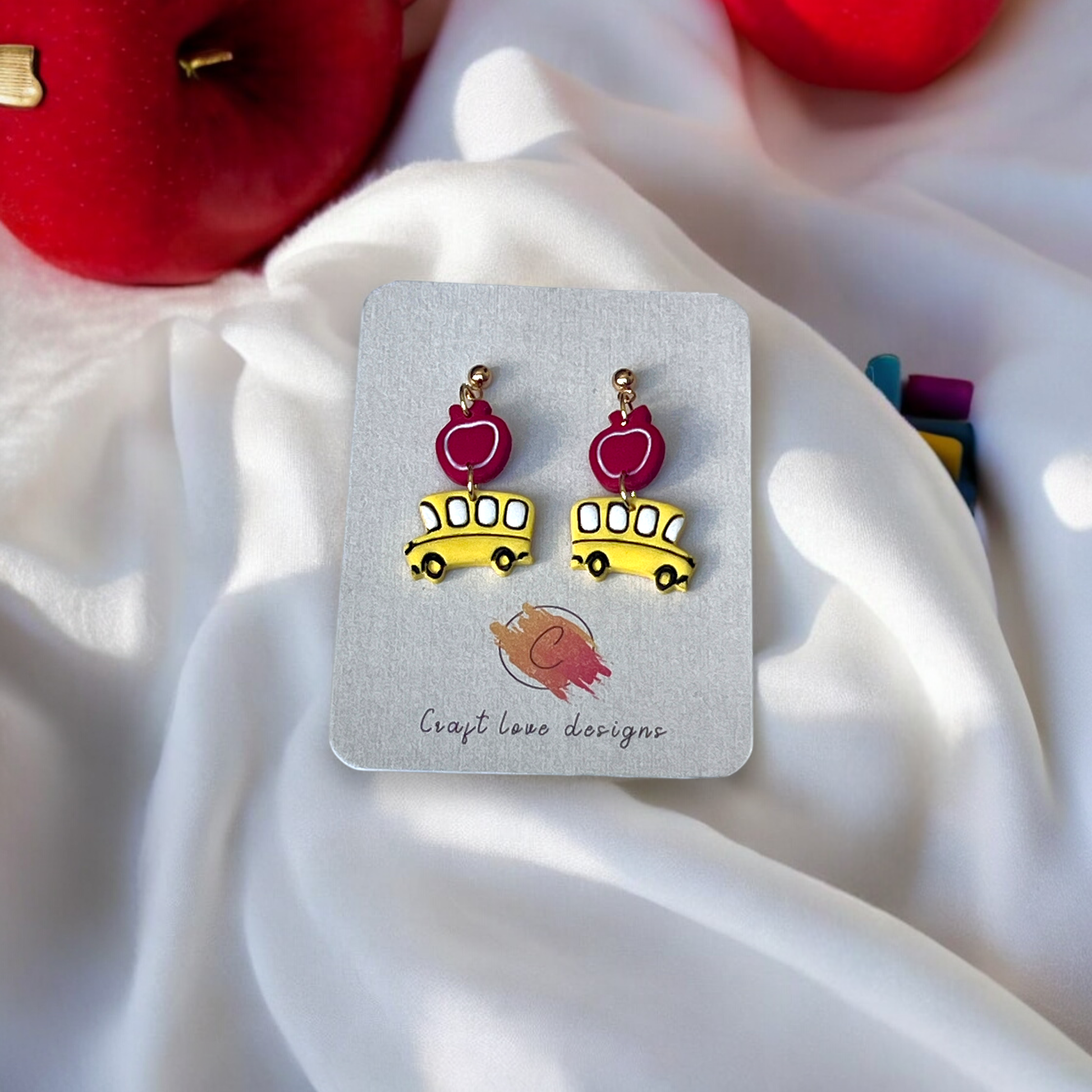 Cute earrings featuring yellow school buses with red apple accents, displayed on a designer card against a soft fabric and apples background.