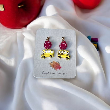 Cute earrings featuring yellow school buses with red apple accents, displayed on a designer card against a soft fabric and apples background.