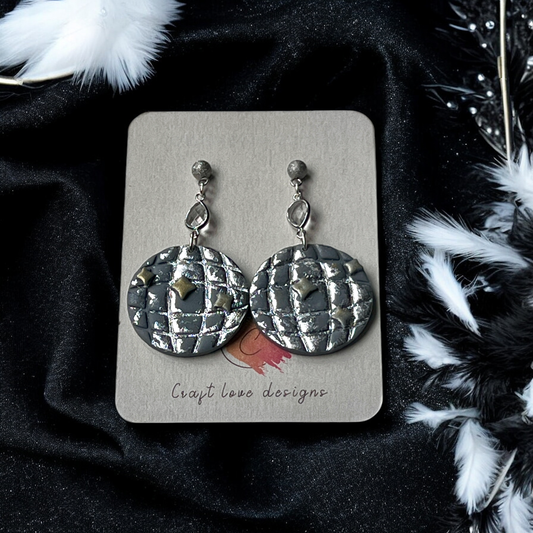 Round earrings with metallic silver texture and star accents, dangling from crystal studs, displayed on a jewelry card with black fabric background.
