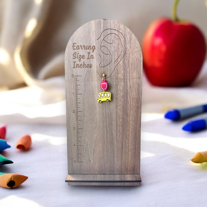 Single school bus and apple-themed earring displayed on a wooden size guide stand, with scattered crayons and a red apple in the background. 