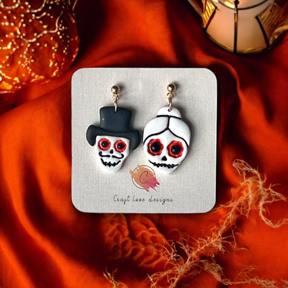 Halloween skeleton couple earrings featuring a skull with a top hat and another with a bun, set on a display card against orange fabric. 