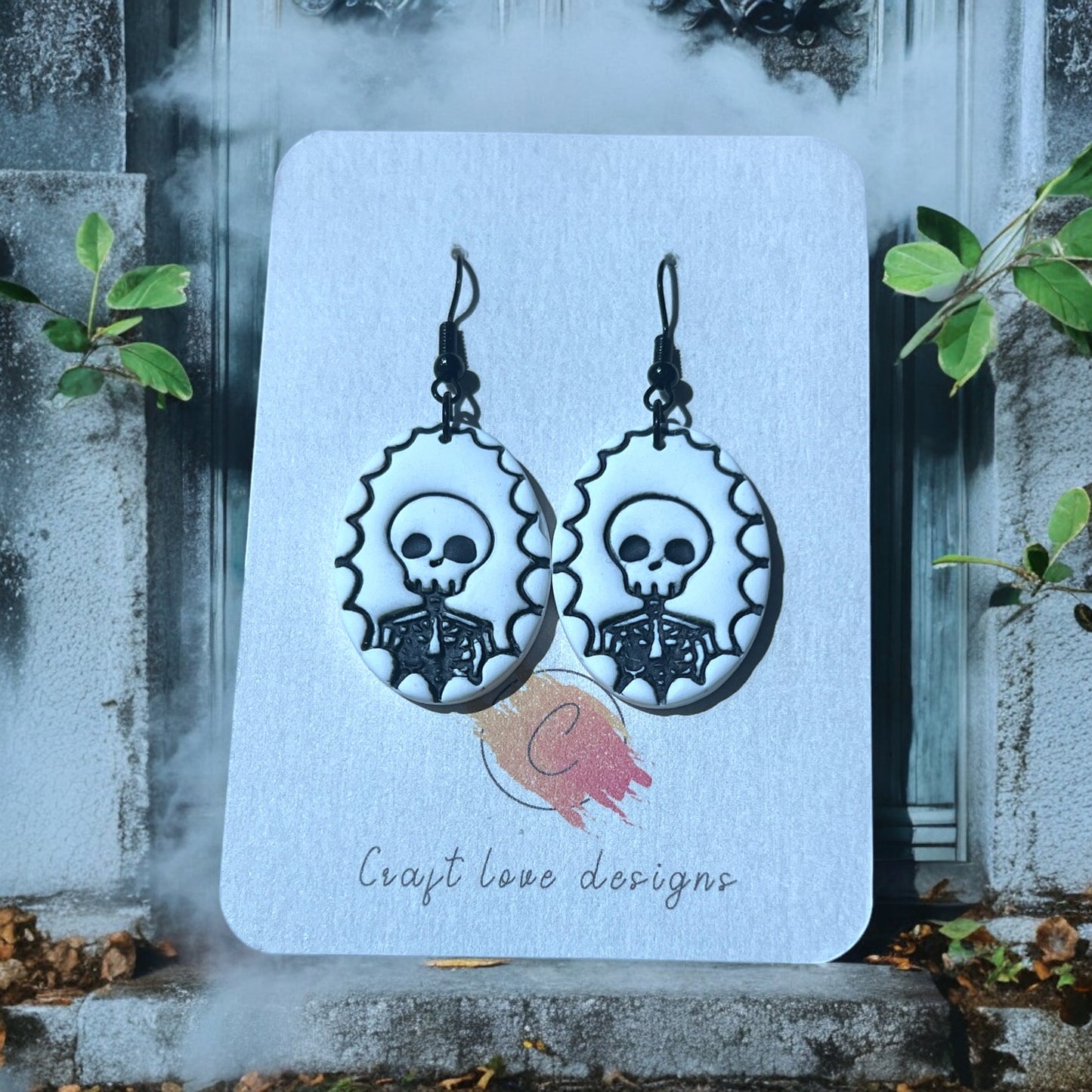 Black and white skeleton cameo earrings on a Craft Love Designs display card, set against a foggy, eerie background with gothic decor. 