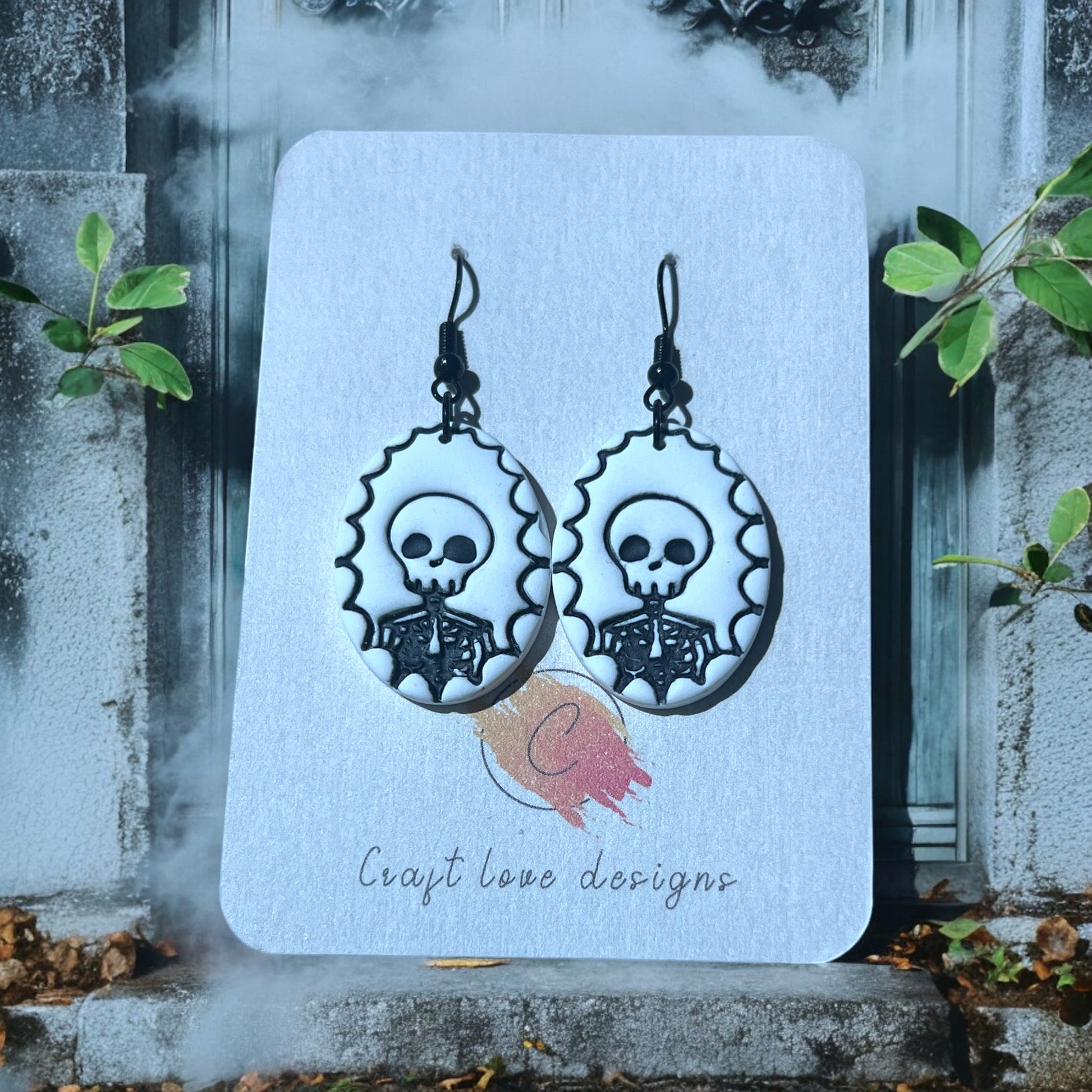 Black and white skeleton cameo earrings on a Craft Love Designs display card, set against a foggy, eerie background with gothic decor. 