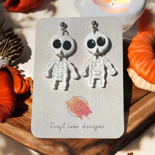 Black and white skeleton cameo earrings on a Craft Love Designs display card, surrounded by pumpkins and set in a spooky, dim setting.
