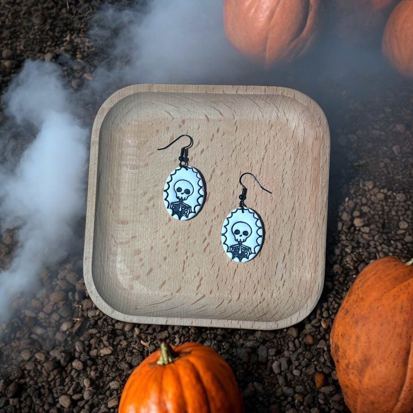 Black and white skeleton cameo earrings resting in a wooden tray, surrounded by pumpkins and fog for a spooky Halloween display. 