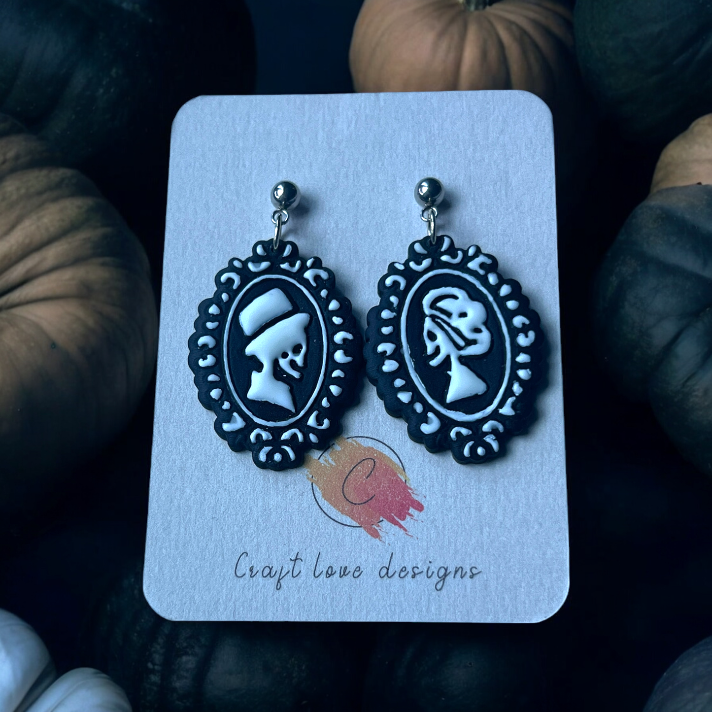  Black and white skeleton cameo earrings on a Craft Love Designs display card, surrounded by pumpkins and set in a spooky, dim setting.