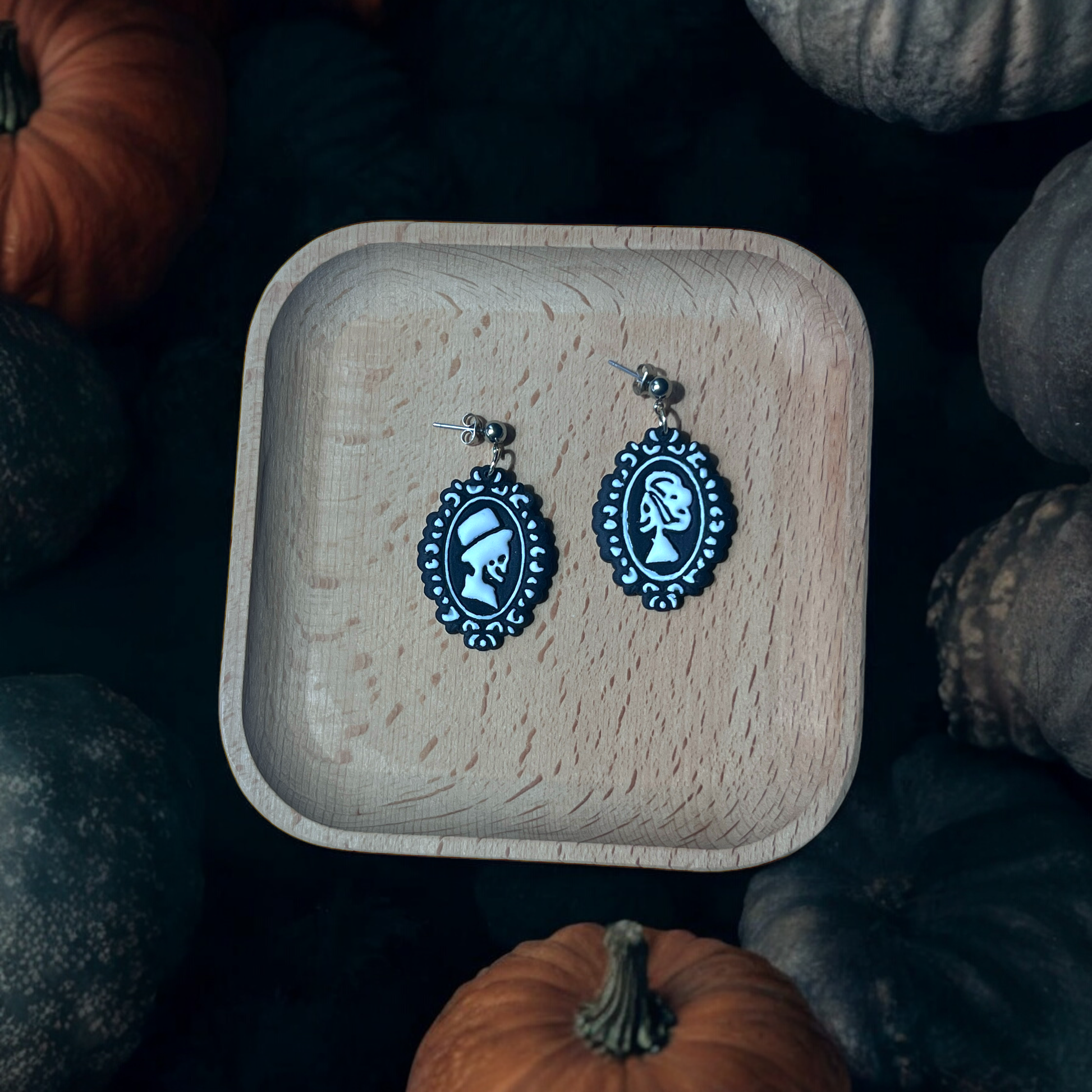 Black and white skeleton cameo earrings resting on a wooden tray, surrounded by autumn pumpkins, creating a festive and spooky display.