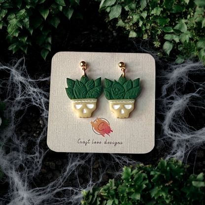 Clay earrings shaped like skull planters with leafy green succulents, displayed on a card amidst spooky cobweb accents and greenery.