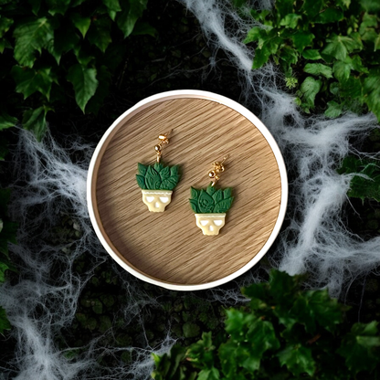 Unique skull-shaped planter earrings topped with textured green leaves, resting on a wooden tray surrounded by cobwebs and foliage. 
