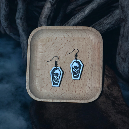 Black and white coffin earrings with skull details resting in a wooden tray, surrounded by foggy decor and eerie wooden textures.