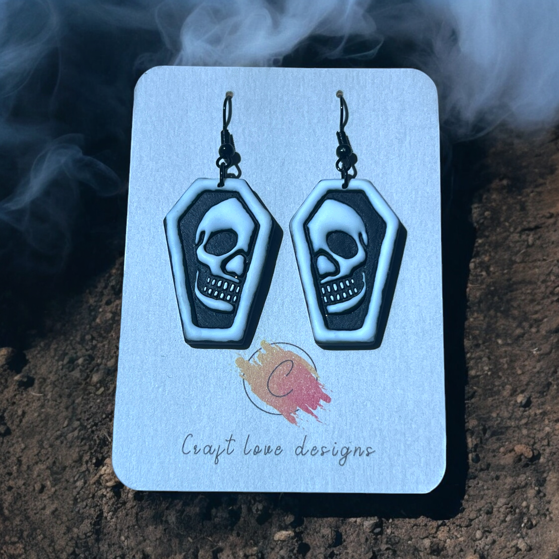 Black and white coffin-shaped earrings with skull designs on a Craft Love Designs display card, set against a foggy, spooky background. (