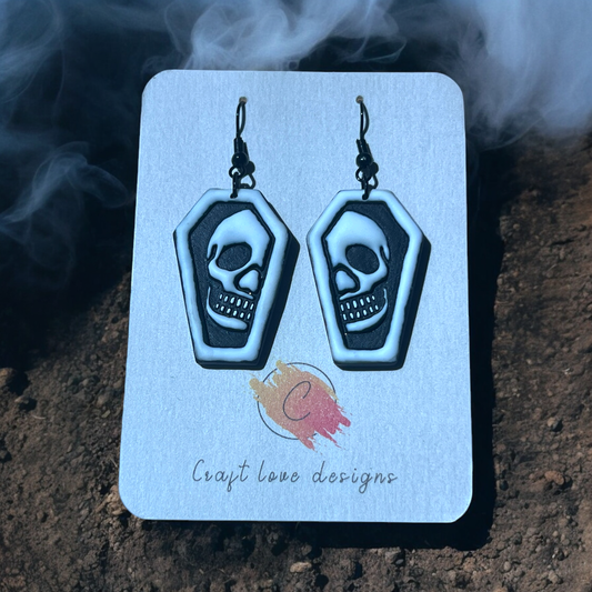 Black and white coffin-shaped earrings with skull designs on a Craft Love Designs display card, set against a foggy, spooky background. (