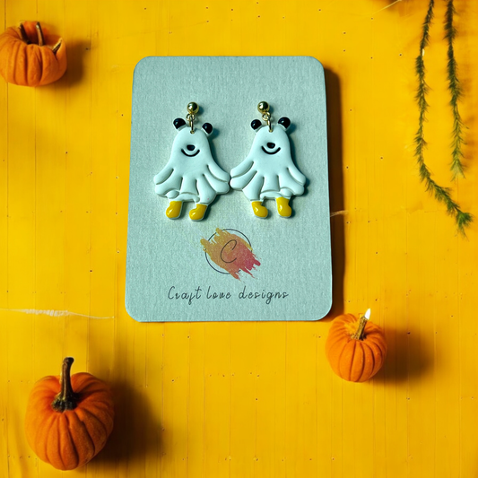 Fun and whimsical ghost earrings with black ears and yellow boots, placed on a branded card with a pumpkin-themed background.