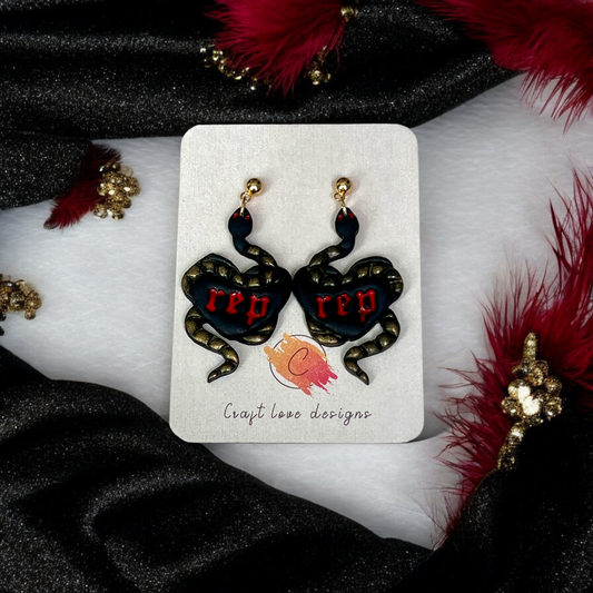 Gold-detailed snake earrings wrapped around black hearts with red “rep” text, displayed on a branded card over black and red fabric.