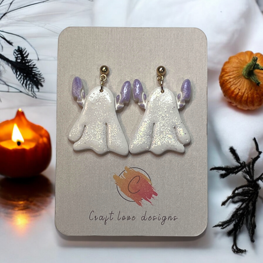 Sparkling white ghost earrings with lavender accents and glitter finish, displayed on a branded earring card among cozy Halloween decor.