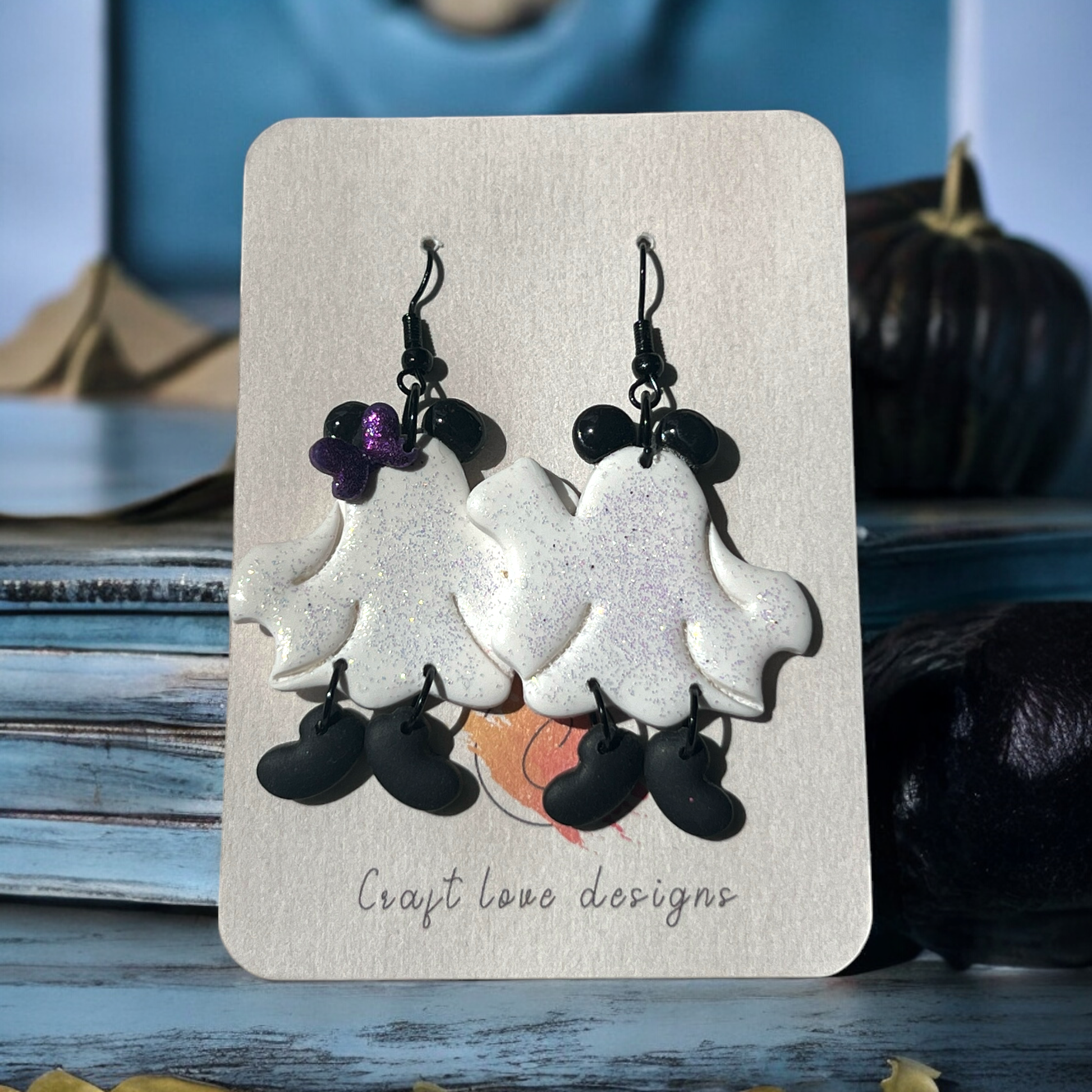 Sparkly white ghost earrings featuring a cute purple bow, dangling black shoes, and a playful Halloween theme on Craft Love Designs backing.