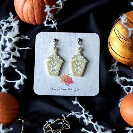 Cream-colored coffee cup earrings with spider web design, displayed on a card surrounded by pumpkins, cobwebs, and a dark Halloween setting.