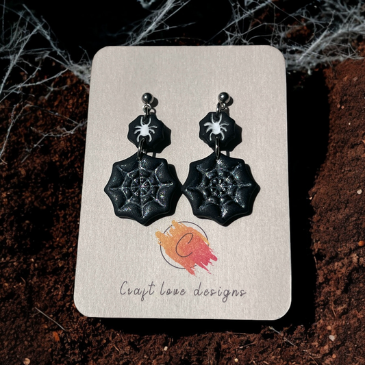 Black glittery spider web earrings with small spider accents on a Craft Love Designs display card, set against dirt and cobwebs. 