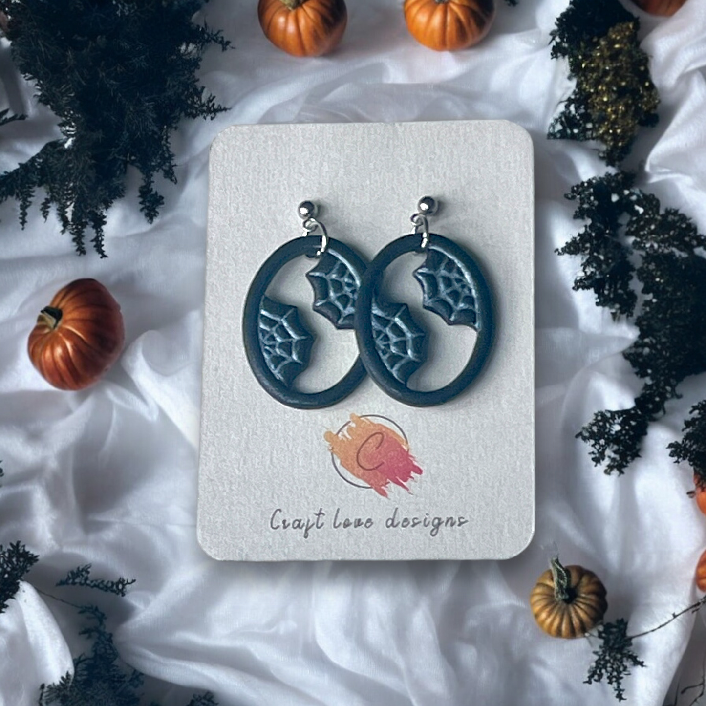 Oval-shaped earrings featuring spider web details on a Craft Love Designs display card, surrounded by mini pumpkins and spooky decor. 
