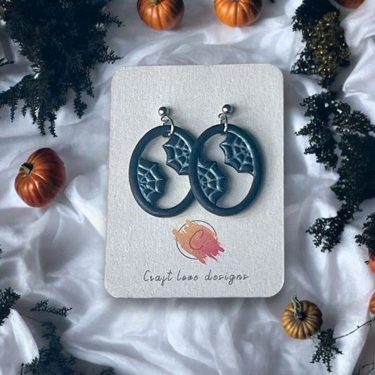 Oval-shaped earrings featuring spider web details on a Craft Love Designs display card, surrounded by mini pumpkins and spooky decor. 