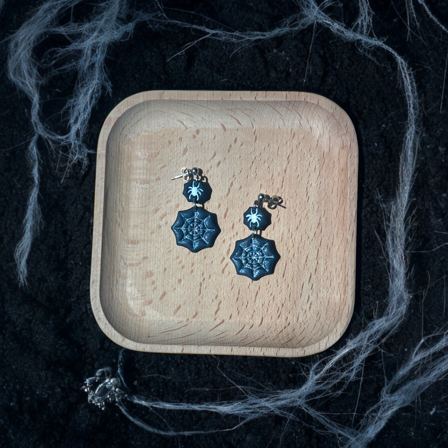 Black spider web earrings with glitter details and small spider charms resting in a wooden tray, surrounded by eerie cobwebs and dark soil. 