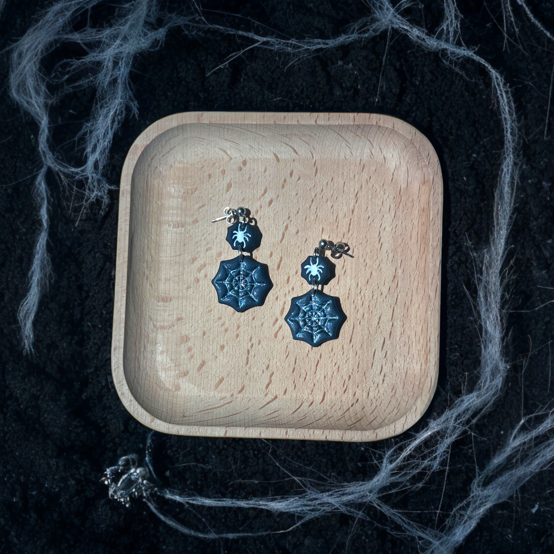 Black spider web earrings with glitter details and small spider charms resting in a wooden tray, surrounded by eerie cobwebs and dark soil. 