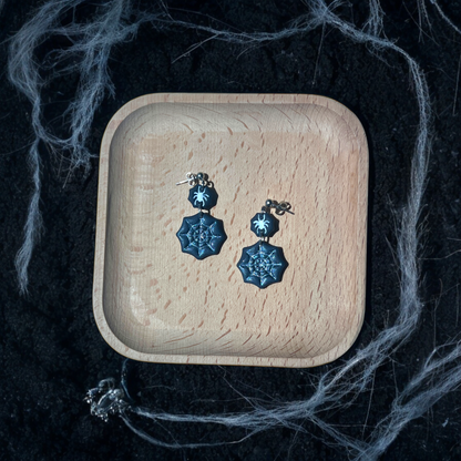 Black spider web earrings with glitter details and small spider charms resting in a wooden tray, surrounded by eerie cobwebs and dark soil. 