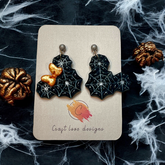 Handmade black spiderweb earrings featuring a sparkly orange bow, displayed on a decorative Halloween-themed card. 