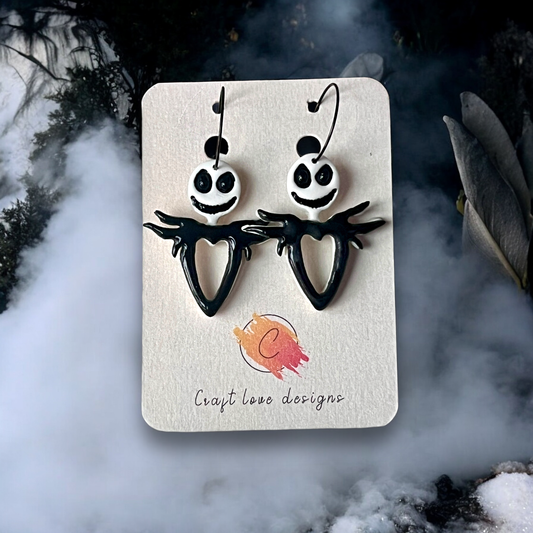 Halloween-themed skeleton dangle earrings with grinning faces and heart-shaped bodies on a display card, surrounded by eerie mist.
