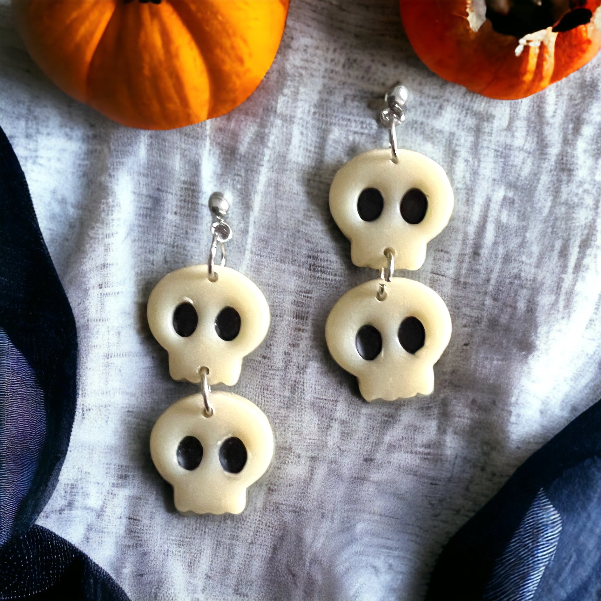 White spooky skull-shaped dangle earrings arranged on a soft fabric with mini pumpkins surrounding the setup.