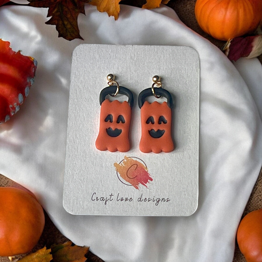 Pumpkin-shaped earrings with "Spooky" text on a Craft Love Designs display card, surrounded by red fabric and festive Halloween decor