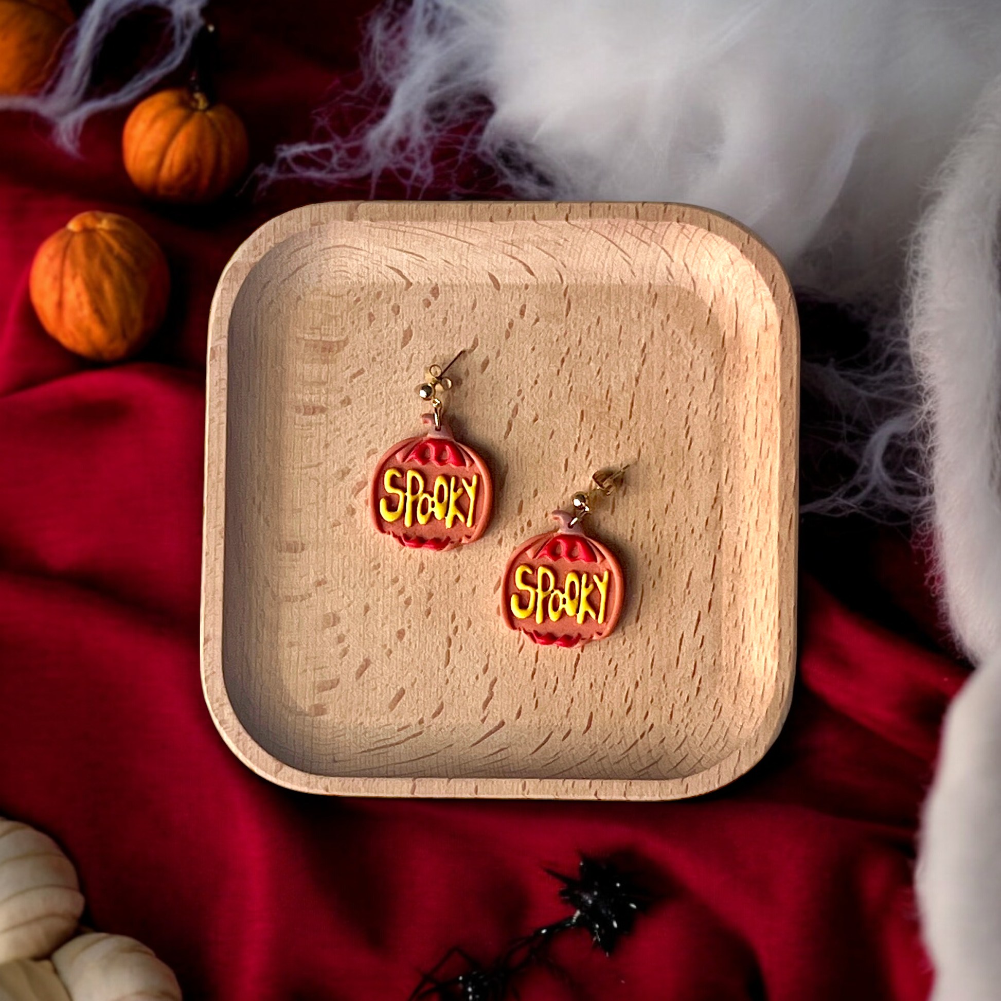 Pumpkin-shaped earrings with yellow "Spooky" text resting in a wooden tray, surrounded by red fabric and spooky Halloween accents. 
