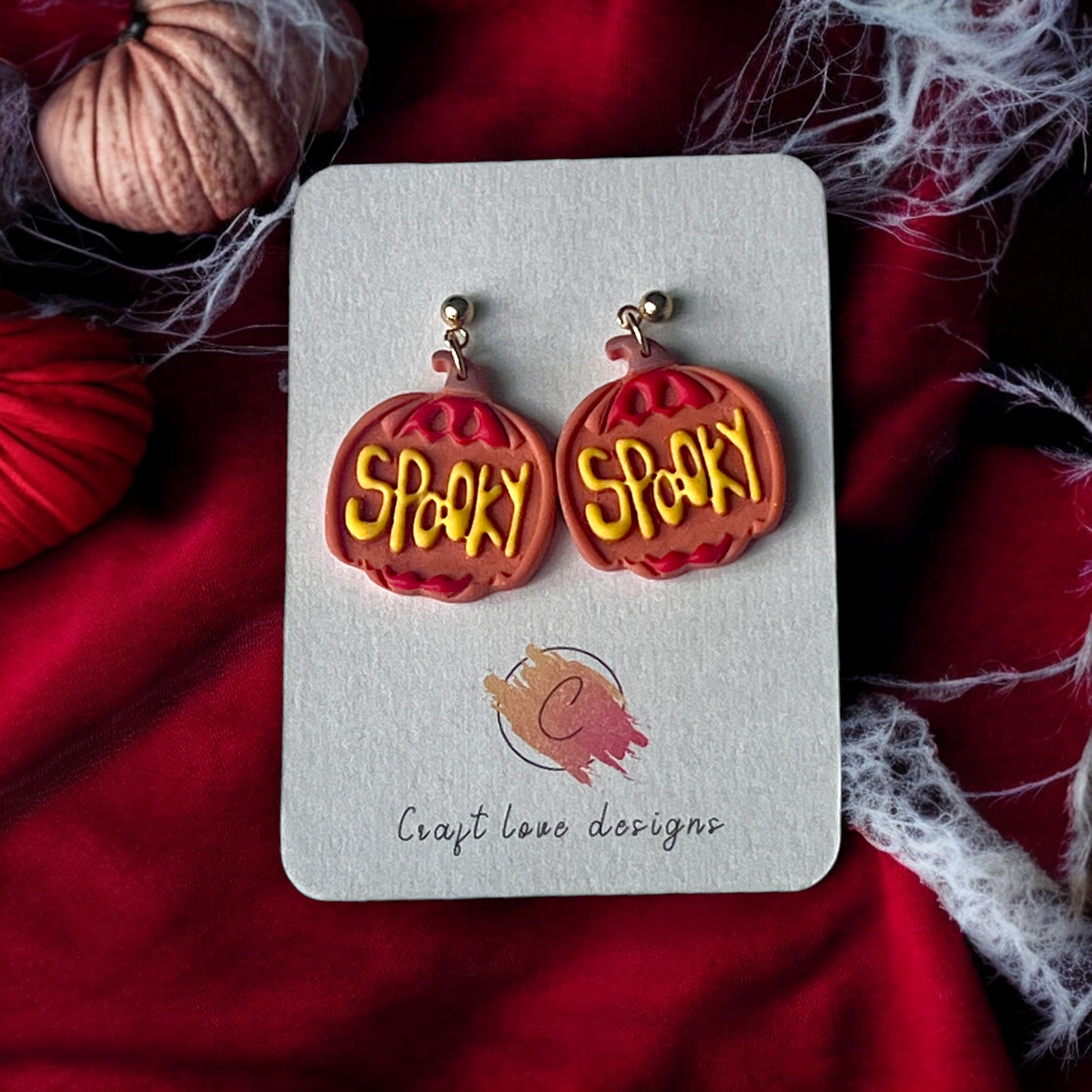 Pumpkin-shaped earrings with "Spooky" text on a Craft Love Designs display card, surrounded by red fabric and festive Halloween decor. (