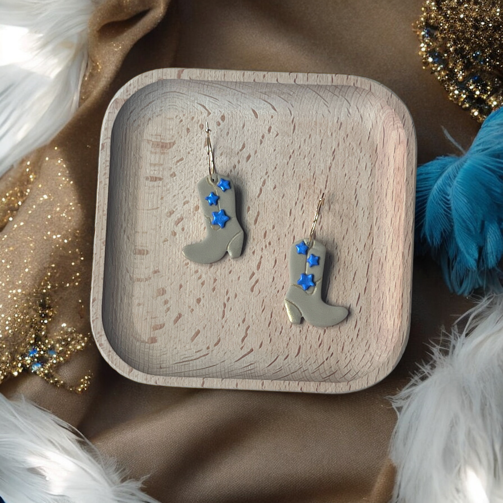 Beige boot-shaped dangle earrings with bright blue stars, placed on a wooden tray, surrounded by soft feathers and shimmering gold fabric.