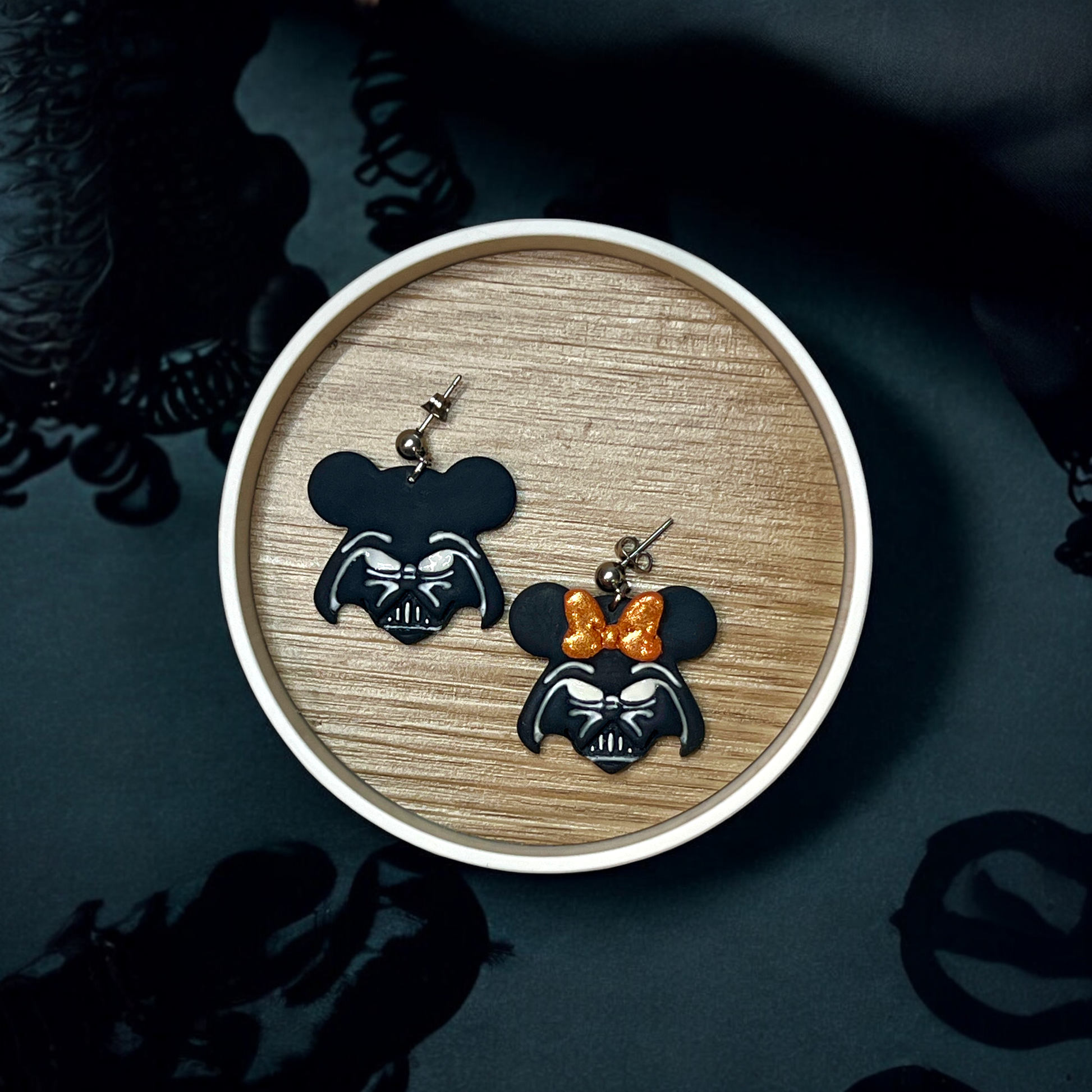 Pair of black mouse-themed earrings featuring a golden glitter bow on one, placed in a round wooden tray with a galactic background. 