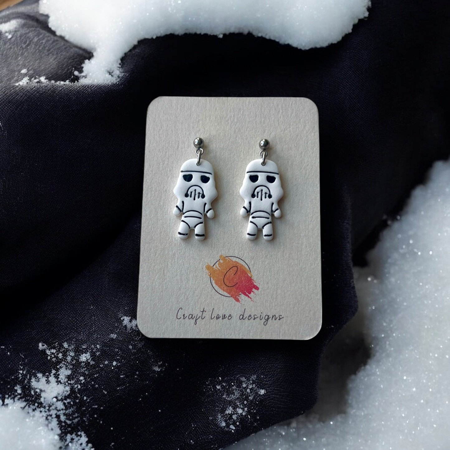 Pair of white stormtrooper-themed earrings displayed on a card, resting on a dark fabric background with scattered snowflakes. 