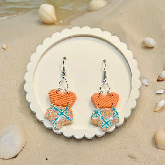 Summer-inspired polymer clay earrings shaped like swimsuits in orange and blue patterns, evoking a playful beachside charm. 